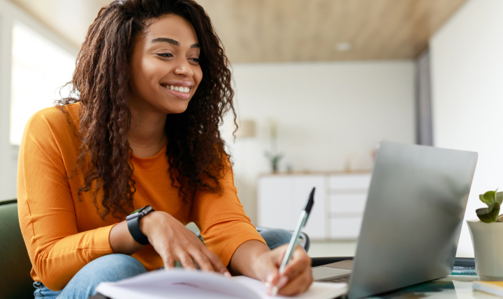 a female student with textured brown hair wearing an orange long sleeve shirt holds a pen while looking at a laptop screen